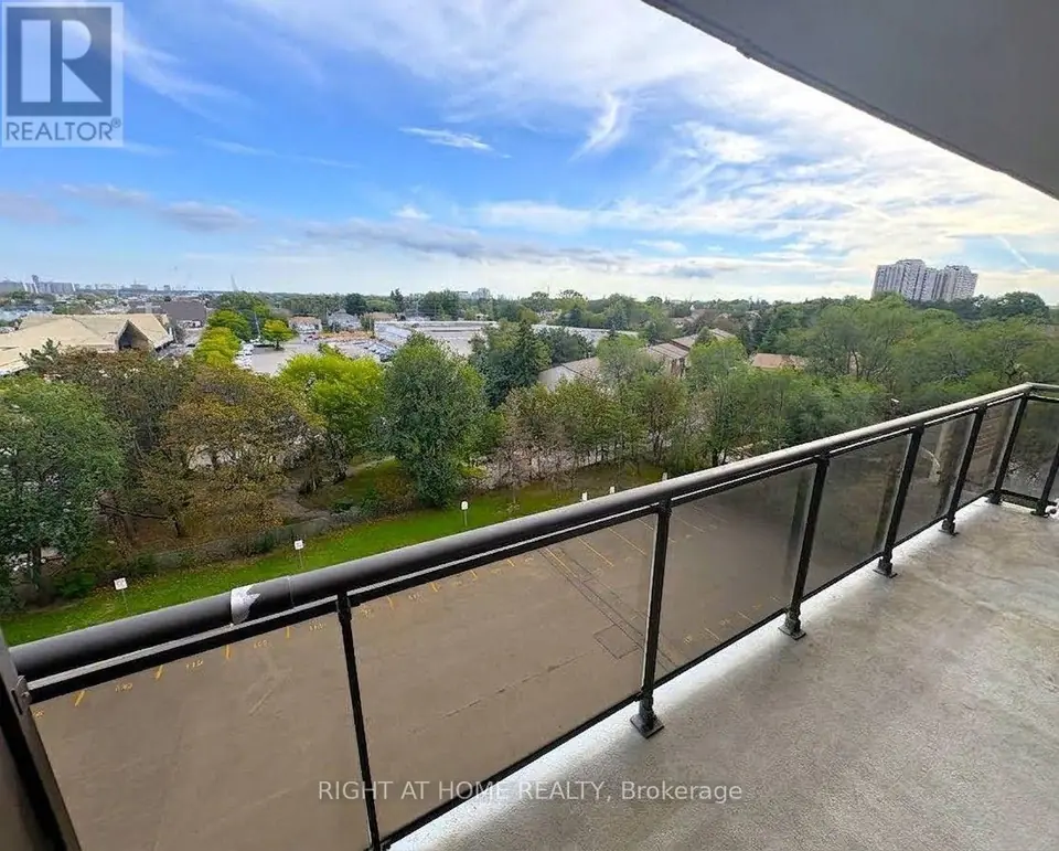 Balcony with glass-panel railing overlooking a parking lot, trees, low-rise buildings and distant high-rise towers under a cloudy sky.