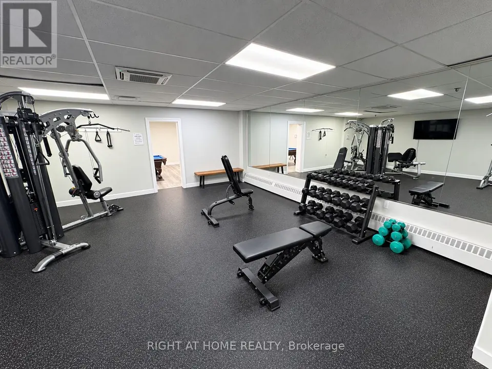 Exercise room with a dumbbell rack, adjustable benches, a cable machine and a wall mirror; a billiards table is visible through the doorway.