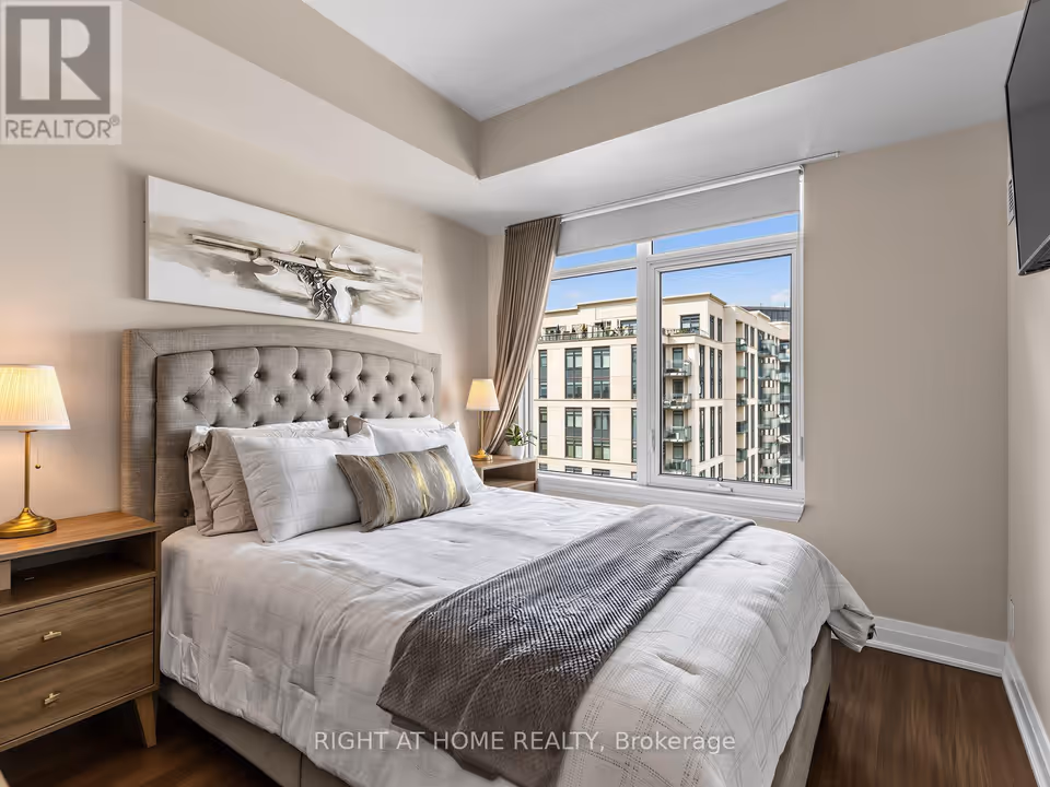A bed with a grey tufted upholstered headboard, white bedding and a grey knit throw sits between two wooden nightstands with brass-toned lamps, below a long abstract painting. To the right a white-framed window with a roller shade and taupe curtain looks across to a neighbouring building with balconies and a rooftop terrace, with a stepped ceiling bulkhead overhead and dark wood-look laminate on the floor.