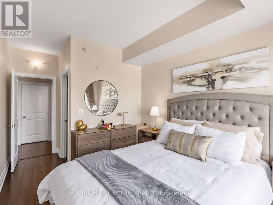 At the left, an open white panel door leads to a short passage with a closed door at its end, next to a second door standing slightly open onto a dark space. To the right, a wooden dresser with a tray of small bottles and a gold heart ornament sits under a round black-framed mirror, beside a bed with a grey tufted headboard, white bedding and an abstract painting above.