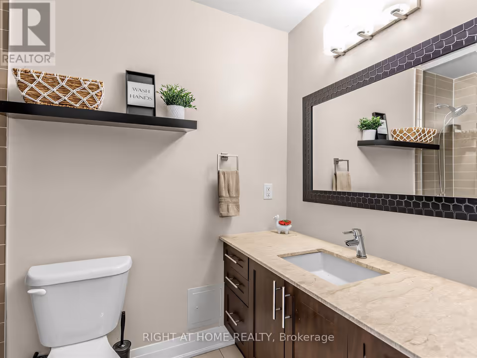 Bathroom vanity wall. A white toilet and a floating black shelf holding a woven basket, a small plant and a “Wash hands” sign sit at left, and a dark brown vanity with a cream, lightly veined counter, undermount sink and chrome faucet runs along the right. A wide mirror with a dark textured frame and a three-light fixture hangs above and reflects the tiled shower.