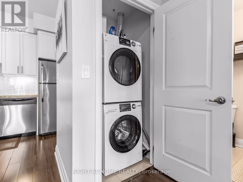 A narrow closet behind an open white panel door holds a stacked washer and dryer, both front-loading, with a foil exhaust duct and a few household items on top. At left are white cabinets, a stainless steel dishwasher and a stainless steel fridge on dark wood-look flooring.