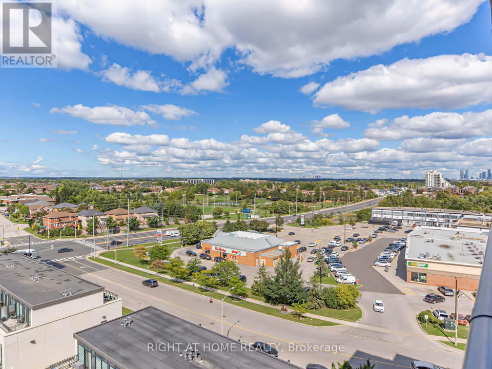 A high-angle view with flat rooftops in the foreground and a wide road running past an orange-brick restaurant with a Tim Hortons sign and a strip plaza with a busy parking lot. Low-rise houses, a green open field and a few distant towers fill the middle distance and horizon beneath a large cloudy sky.