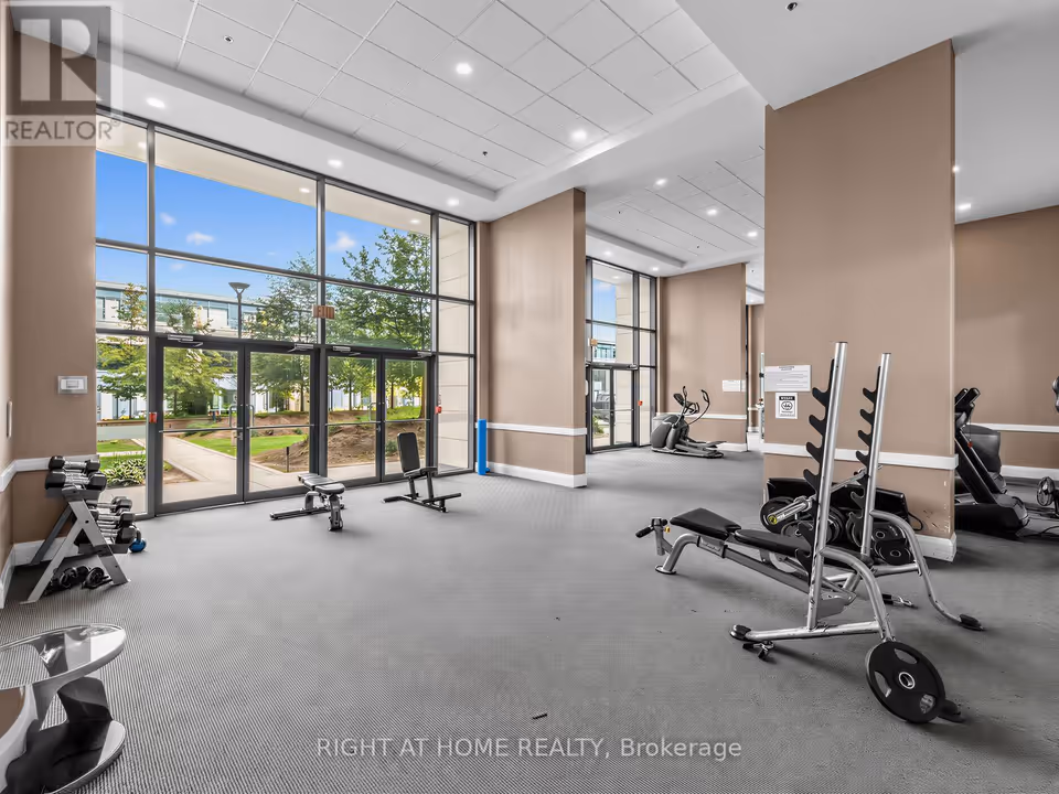 Wide view of a gym with grey carpet and beige columns, looking toward a tall glass wall and double glass doors that face a landscaped path and trees. Dumbbells on a rack, two adjustable benches and a weight rack with plates sit in the foreground, with cardio machines along the right and back.
