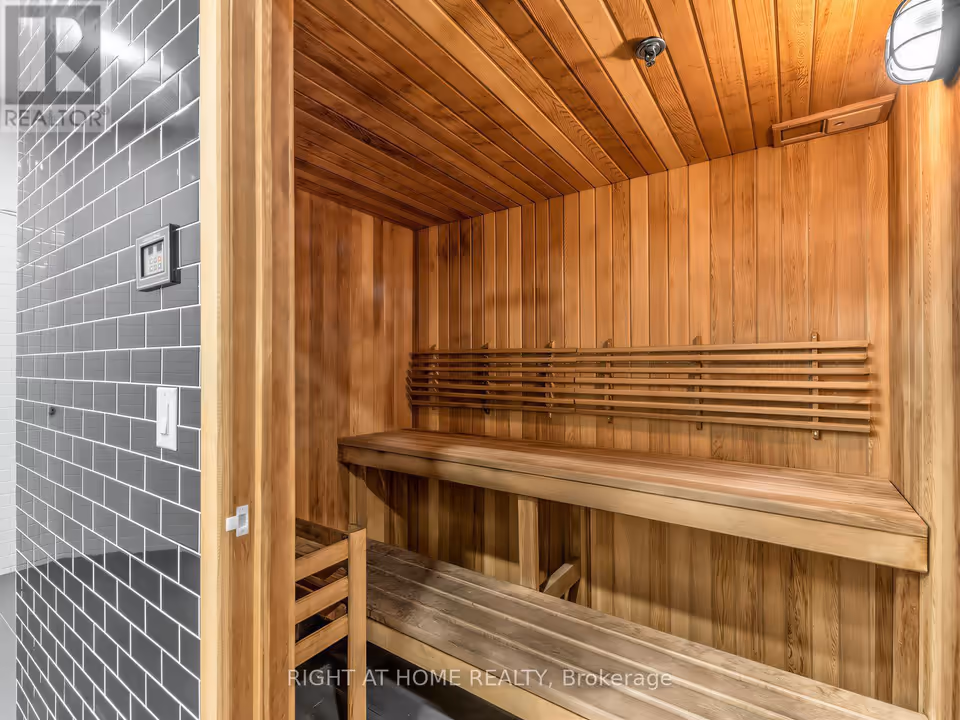 Interior of a sauna lined in light wood, seen from the doorway, with a lower and an upper bench along the back wall, a slatted backrest above them, a sprinkler head in the ceiling and a caged light at top right. At left, a control panel and a light switch are mounted on grey subway tile.